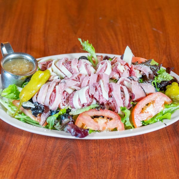 A large Italian salad with ham, cheese, tomatoes, olives, and a side of dressing.