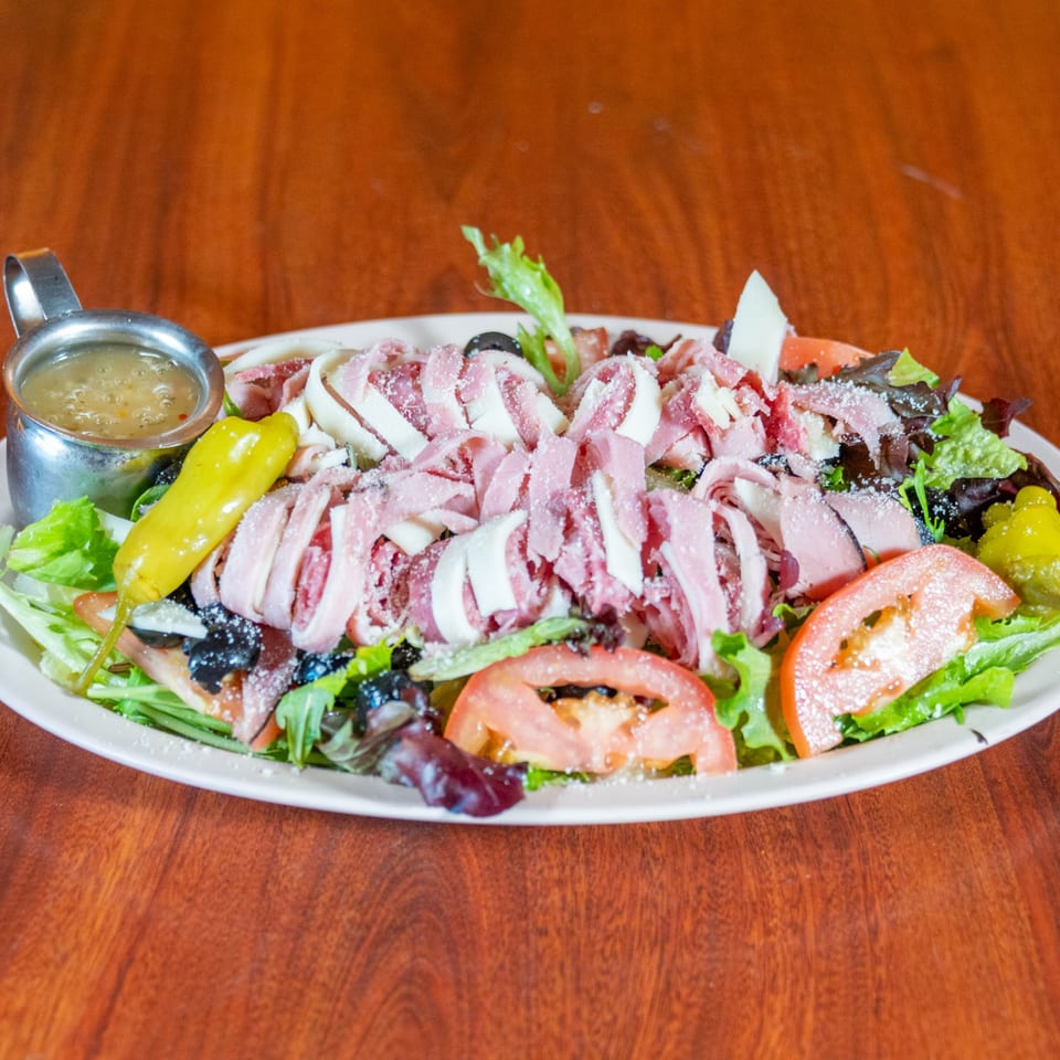 A large Italian salad with ham, cheese, tomatoes, olives, and a side of dressing.