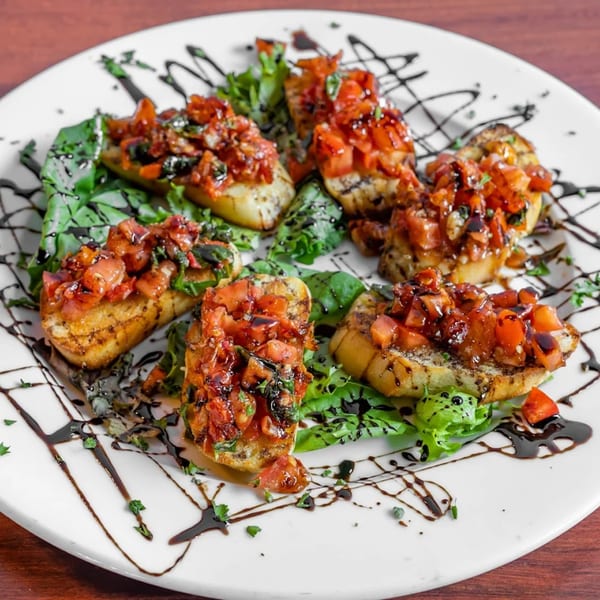 Six bruschetta pieces with diced tomatoes, herbs, and balsamic glaze on a white plate with greens.