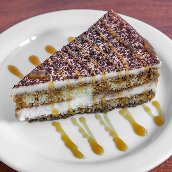 Slice of tiramisu cake with chocolate dusting and caramel drizzle on a white plate.